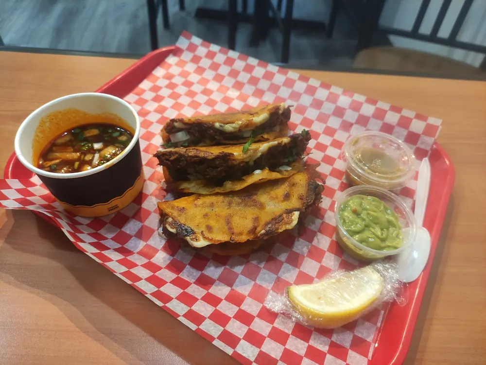Three stacked griddled birria tacos with melted cheese on a red tray with a cup of consomm&eacute;, guacamole and salsa