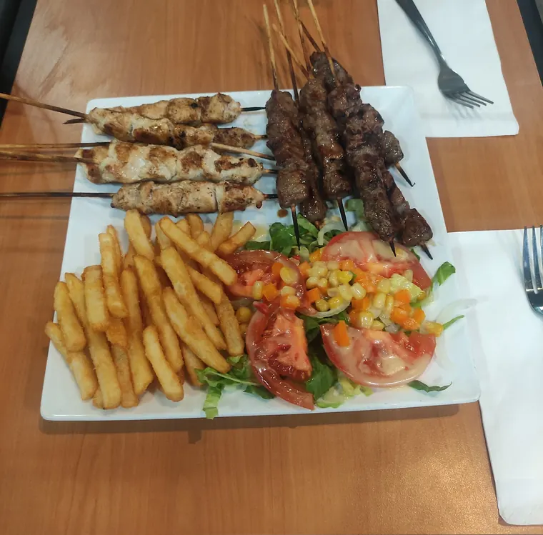 A white plate of grilled chicken and beef skewers with fries and a tomato salad