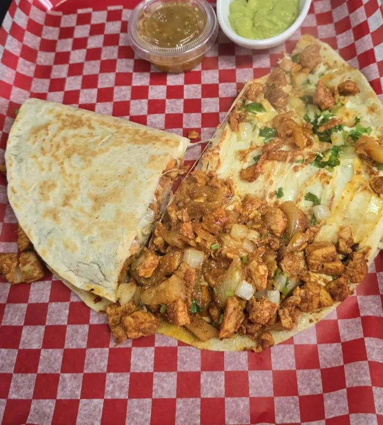 A flat chicken quesadilla cut in halves with guacamole and salsa on checkered paper